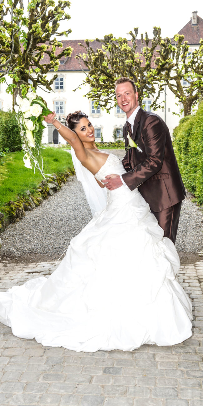 Glückliches Brautpaar in eleganter Pose, die Braut strahlt und hebt ihren Brautstrauß, während der Bräutigam sie liebevoll hält, vor malerischer Schlosskulisse.