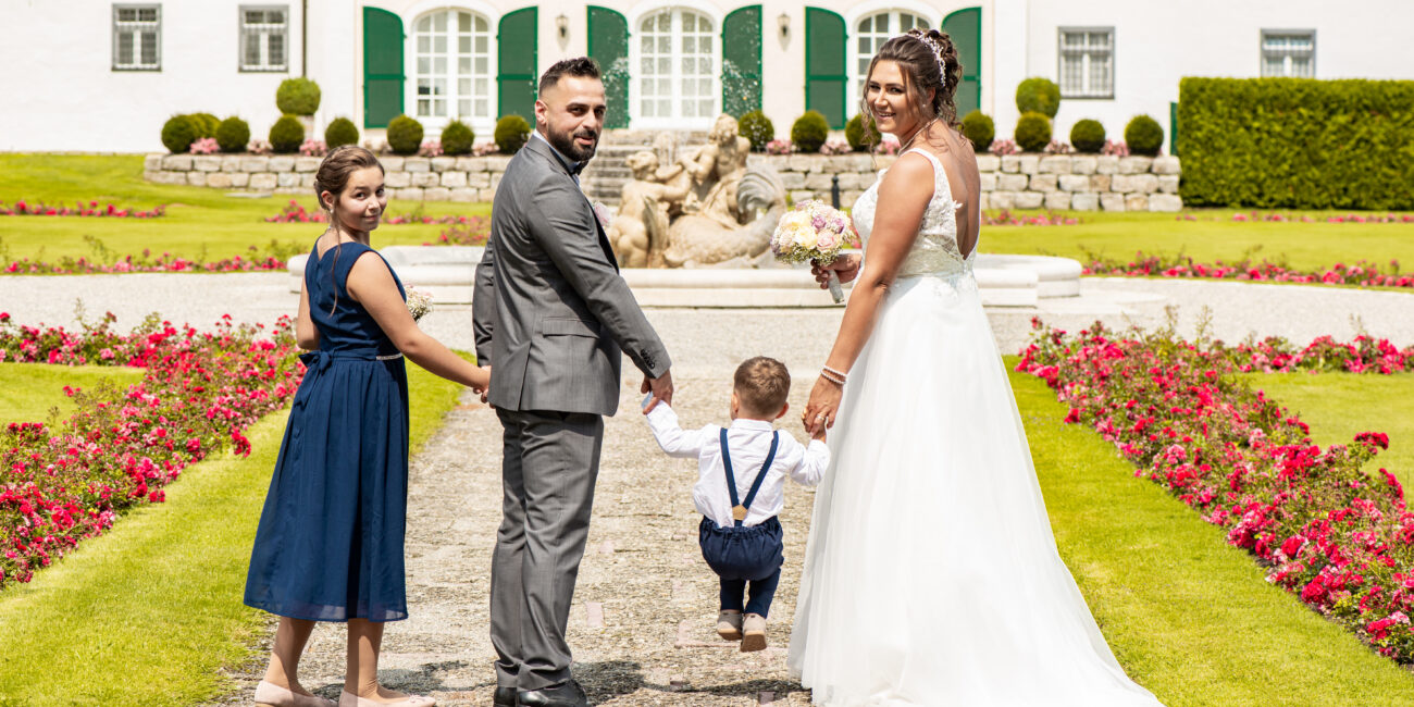 Die Hochzeitsgesellschaft spaziert Hand in Hand durch einen eleganten Garten, mit einem Schloss im Hintergrund. Ein idyllischer Moment voller Freude und Verbundenheit.