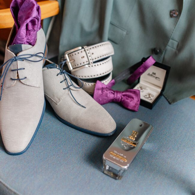 Detailaufnahme der Hochzeitsaccessoires des Bräutigams mit eleganten Schuhen, Gürtel, Fliege und Manschettenknöpfen auf einem Stuhl arrangiert.